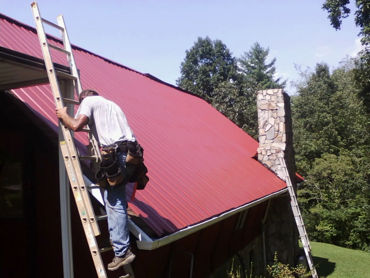 Roofer on a ladder beside a red metal roof and stone chimney — Soffit & Fascia Repair by Roofing Brothers in Broad Run